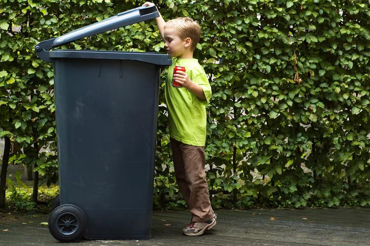 Ein Junge wirft eine leere Getränkedose in eine große Mülltonne im Garten, die von einer Hecke umgeben ist. Eine Mülltonnenbox 4er könnte diese Mülltonne elegant verstecken und den Garten aufgeräumter wirken lassen.