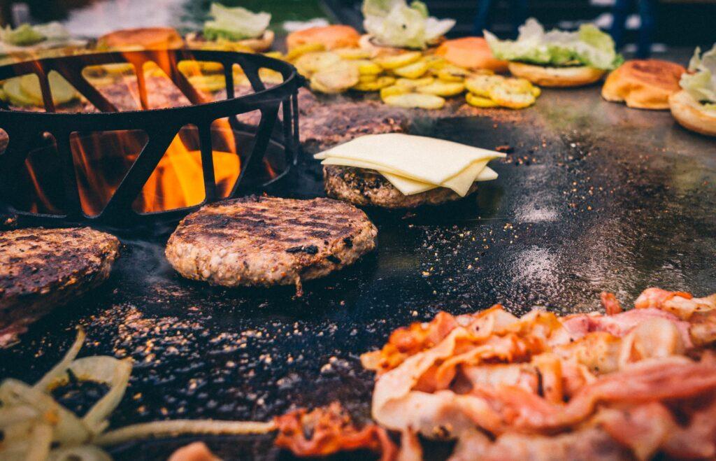 Burger und Bacon brutzeln auf einer Feuerplatte, während Flammen im Hintergrund lodern. Auf der Grillplatte werden Burgerpatties, Käse und weitere Zutaten zubereitet.