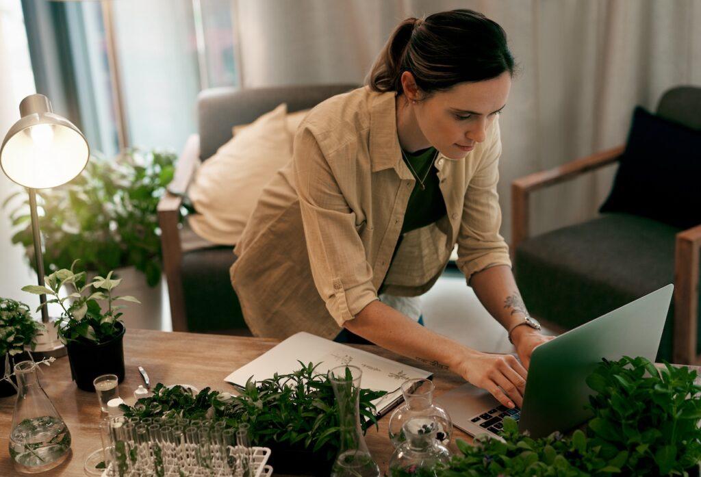 Frau arbeitet an einem Laptop in einem Homeoffice mit Pflanzen und Laborglas, umgeben von nachhaltigen Projekten und botanischen Experimenten