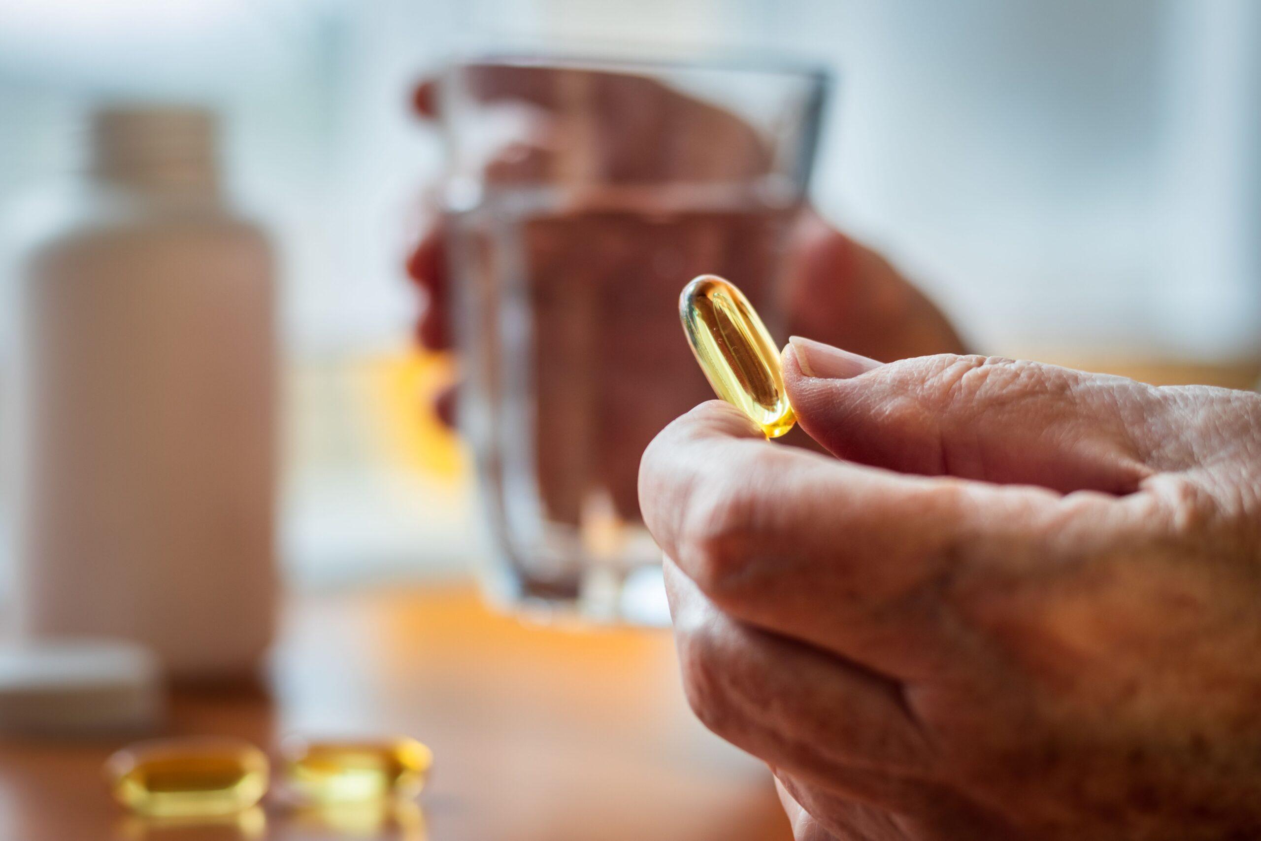 Senior hält eine Kapsel mit Omega-3 Algenöl in der Hand, daneben ein Glas Wasser
